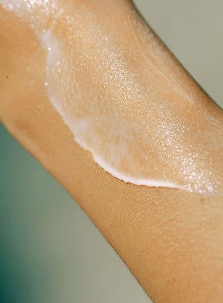 A close-up of a person’s arm with a streak of hydrating cream applied to the skin, giving it a shiny and moisturized appearance. Skin tone is light to medium. The cream is white with a semi-gloss finish and has been spread in one broad line along the forearm. No product packaging or label text is visible. Background is neutral and out of focus. No other objects present.