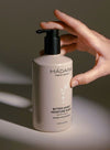 A hand reaches for a beige, matte-finish pump bottle with rounded edges, displaying the text “MÁDARA organic skincare, Bitter Honey Moisture Wash” in black sans-serif font on the front. The product is set against a plain, neutral background. Only the hand and forearm are visible; no other details or props appear in the shot.