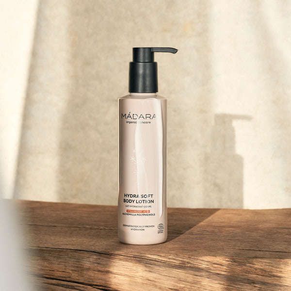 Madara Hydra Soft Vartalovoide; pullo MÁDARA Hydra Soft Body Lotion -vartalovoidetta mustalla pumpulla, pystyssä vaalealla puisella pinnalla; pullo sylinterinmuotoinen mattapinnalla, lihavoitu groteskityyppografia päätekstille; tausta pelkistetty ja neutraali; pehmeä auringonvalo luo varjoja yhdeltä puolelta; ei ihmisiä tai käsiä kuvassa.