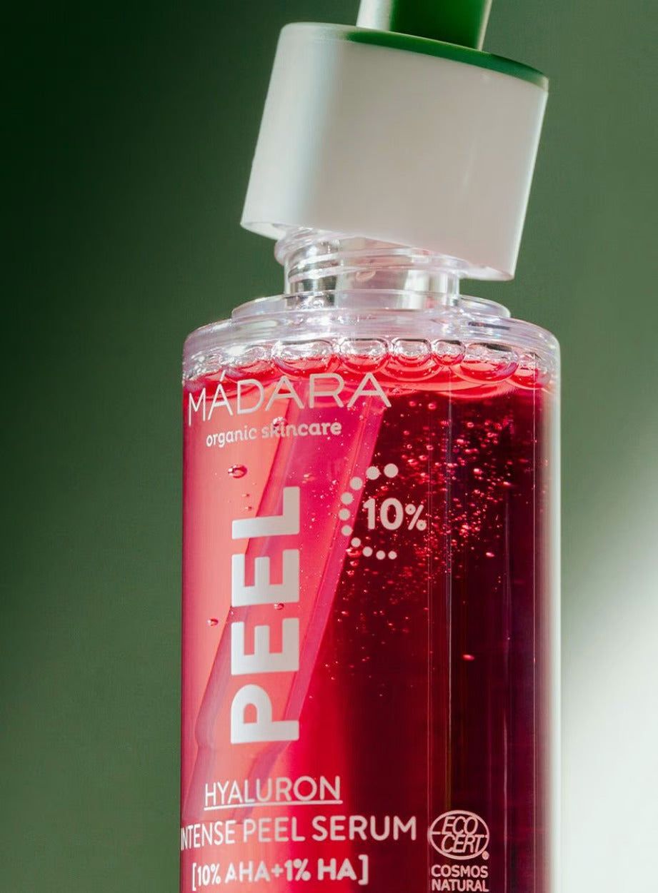Close-up of a bottle of MADARA organic skincare Hyaluron Intense Peel Serum with red liquid and visible bubbles inside. The container is cylindrical, transparent glass with a glossy surface, fitted with a white dropper cap. Label has clean, bold white sans serif text showing product name and brand. Plain light background. No people or hands in frame. Close-up of a bottle of MADARA organic skincare Hyaluron Intense Peel Serum with red liquid and bubbles inside, featuring a dropper cap and white label text.