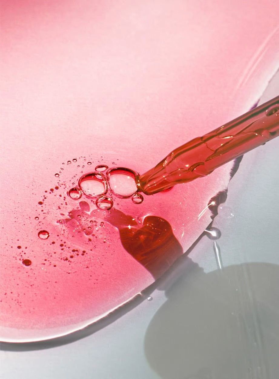 A dropper releases red liquid onto a pink surface, forming bubbles and a glossy texture enhanced by dynamic light and shadow. The liquid is deep red and semi-transparent. The dropper is clear plastic with no visible label or text. No product packaging, hands, or people are present. No background objects are visible; the shot is a close-up above the surface.
