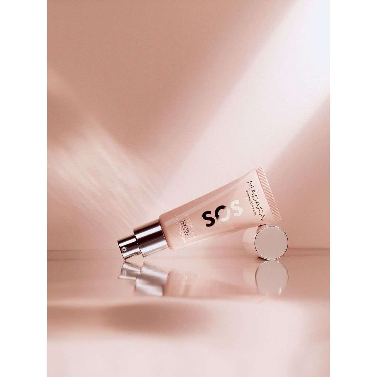 MÁDARA SOS Hydra Recharge Cream -putkilo kyljellään, korkki irti, heijastuu kiiltävältä vaaleanpunaiselta pinnalta lämpimässä, pehmeässä valaistuksessa; putkilo lieriömäinen, muovia, matta pinta; etiketissä tuotenimi yksinkertaisella groteskityypillä; ei ihmisiä tai käsiä kuvassa.