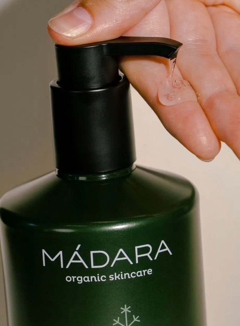 A hand dispenses clear gel from a dark green MÁDARA organic skincare bottle with a black pump top. The cylindrical bottle is opaque and has a matte finish. White sans-serif font displays “MÁDARA” vertically; other product information appears in smaller type below. Background is plain white, close-up shot focuses on the bottle and hand, with no other objects visible.
