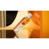 Mossa Vitamin C Glow Perfecting Toner; lieriömäinen muovipullo kallellaan kiiltävää oranssia pintaa vasten, alla näkyvä heijastus; valkoinen korkki, oranssia ja harmaata groteskitekstiä painettuna; tausta kirkkaan oranssi terävin mustin ja beigein kaarevin muodoin; ei ihmisiä tai käsiä.