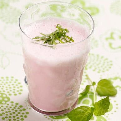 Nutrilett VLCD Mansikkapirtelö; lasi vaaleanpunaista smoothieta silputun vihreän yrtin peittämänä, kuvioidulla valkois-vihreällä alustalla tuoreiden minttulehtien ja eloisien yrttien vierellä; kirkas, lieriön muotoinen lasi ilman brändäystä tai etikettejä; alusta toistuvaa geometrista kuviota valkoisin ja vihrein sävyin; ei ihmisiä tai käsiä.