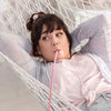 Woman in a light pink top relaxes on a white hammock, sipping through a long pink straw with her arms resting behind her head, enjoying a tranquil moment. White fabric hammock with netted edges; open outdoor setting with greenery visible in background; no packaging or visible labels present. Shot is medium distance showing upper body and surroundings clearly.