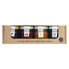 A set of four Finnish jam jars in a cardboard box, each labeled with different berry flavors and vibrant colors—green, red, orange, and blue. Authentic Finnish text appears on both the box and jar labels. The jars are clear glass with metallic lids, cylindrical in shape. Labels have bold printed Finnish words and simple graphics. Cardboard packaging has a matte finish. Jars are upright in the box against a neutral background; no people or hands visible.