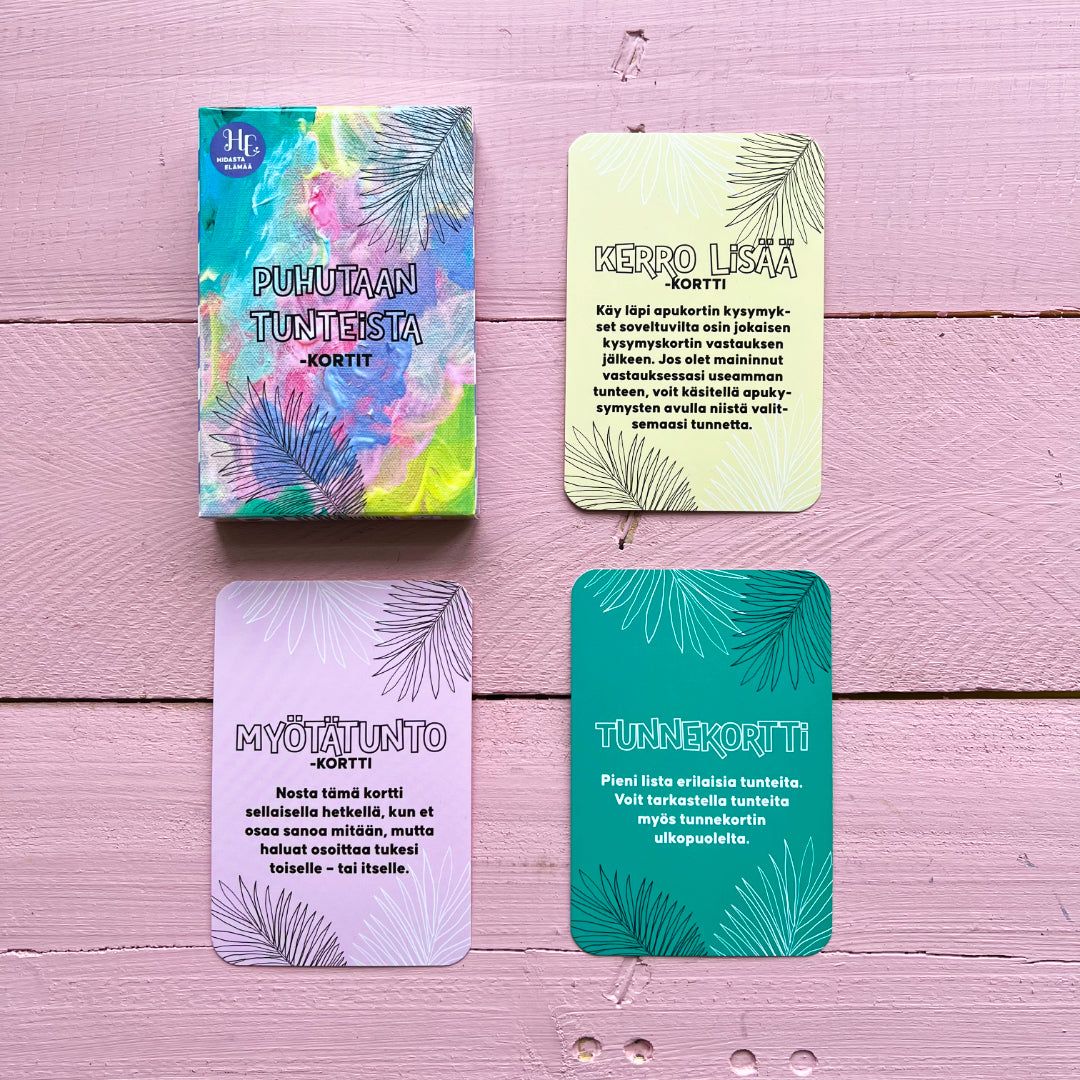 A vibrant card deck box with three emotion-themed Finnish cards displayed on a pink wooden surface. The rectangular box and cards are made of matte cardstock, each card showing distinct text in bold, sans-serif type and featuring a multicolored decorative leaf motif. No people or hands visible in the image. Cards are arranged face-up beside the upright box.
