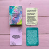 Four emotion-themed rectangular cards are laid out on a matte pink wooden surface. The main card is multicolored with bold sans-serif Finnish text stating “Puhutaan tunteista – kortit.” Three other cards have light pastel backgrounds with subtle leaf motifs and additional Finnish phrases. No packaging, people, or hands are present. Overhead shot, no reflections or glossiness visible.