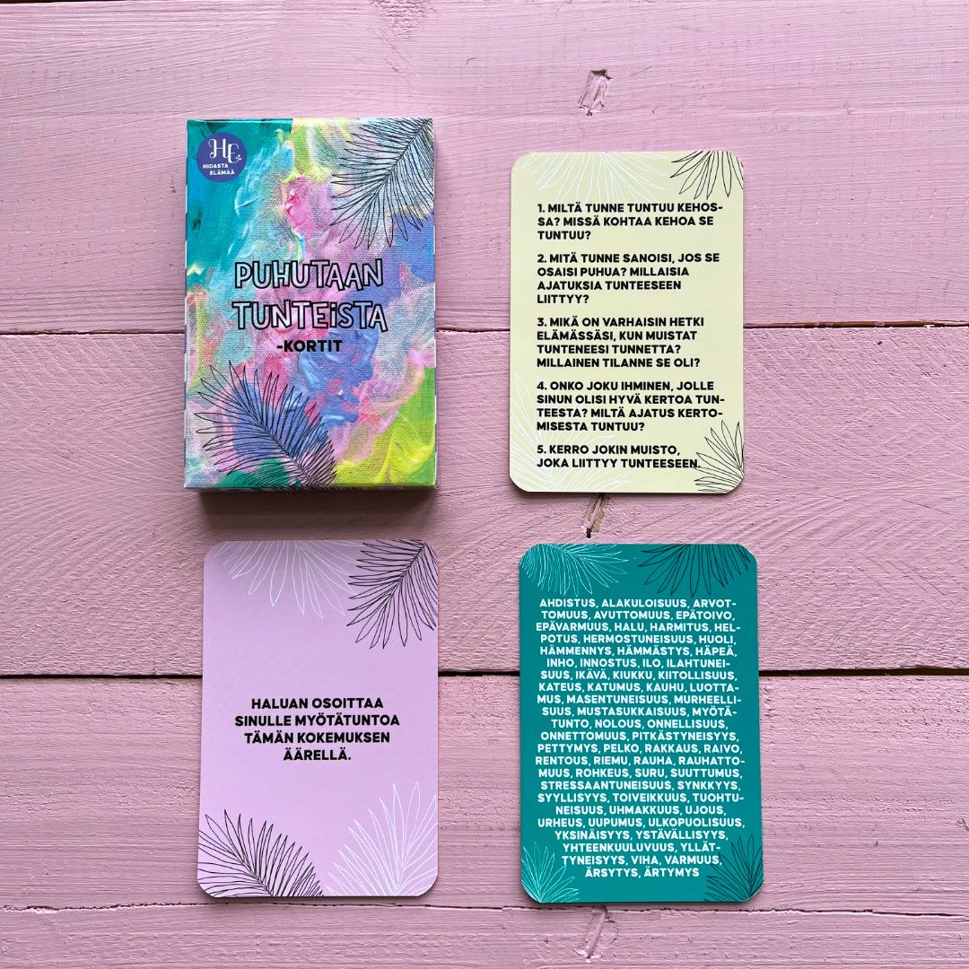 Four emotion-themed rectangular cards are laid out on a matte pink wooden surface. The main card is multicolored with bold sans-serif Finnish text stating “Puhutaan tunteista – kortit.” Three other cards have light pastel backgrounds with subtle leaf motifs and additional Finnish phrases. No packaging, people, or hands are present. Overhead shot, no reflections or glossiness visible.