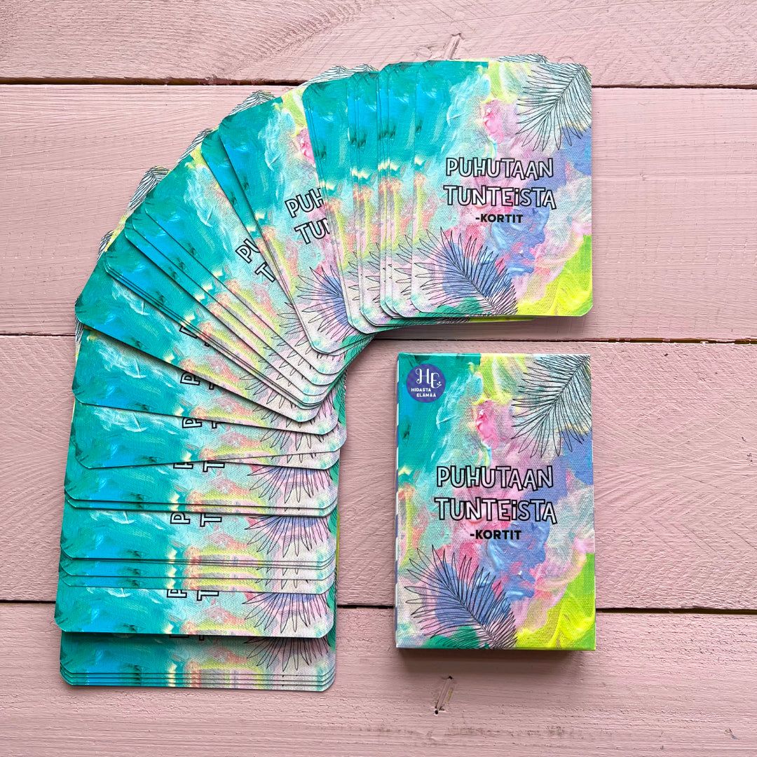 A deck of colorful, rectangular cards with the Finnish text PUHUTAAN TUNTEISTA - KORTIT is displayed on a matte pink wooden surface. The cards have varied solid colors and are fanned out in an arc. One stack is face up on the right, revealing printed text. No packaging or people are visible. A deck of colorful cards with Finnish text PUHUTAAN TUNTEISTA - KORTIT on a pink wooden surface. The emotion-themed cards are fanned out, with one stack shown face up on the right.