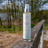 A cylindrical plastic spray bottle labeled “Saaren Taika Merisuola Suihke” stands on a horizontal wooden railing outside. The bottle is white with a clear cap, matte finish, and modern sans-serif typography in blue and gray. No people or hands are present. Background includes green trees, grass, and reflective water under daylight.