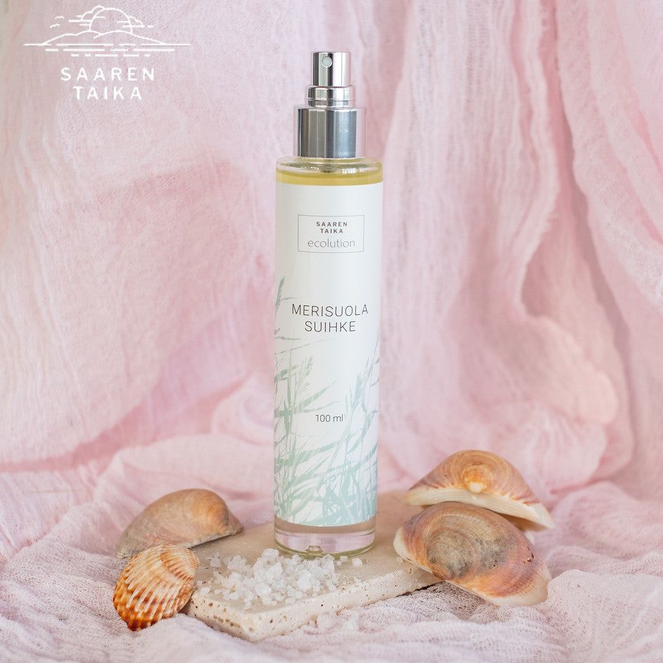 A cylindrical plastic spray bottle labeled “Saaren Taika Merisuola Suihke” stands upright on a flat, light-colored stone slab. Coarse sea salt crystals and two seashells lie nearby on the stone surface. The background is soft pink fabric with gentle folds. No hands or people are present.