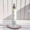 A bottle of Saaren Taika Ecolution Merisuola Sea Salt Spray stands on a white surface with its cap off, in front of a light-striped background. The bottle is cylindrical and white with a matte finish, featuring a silver spray nozzle. Blue, sans-serif text on the label reads “Saaren Taika Ecolution Merisuola Sea Salt Spray.” No people or hands present.