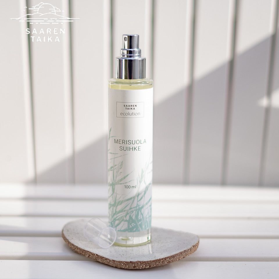 A bottle of Saaren Taika Ecolution Merisuola Sea Salt Spray stands on a white surface with its cap off, in front of a light-striped background. The bottle is cylindrical and white with a matte finish, featuring a silver spray nozzle. Blue, sans-serif text on the label reads “Saaren Taika Ecolution Merisuola Sea Salt Spray.” No people or hands present.