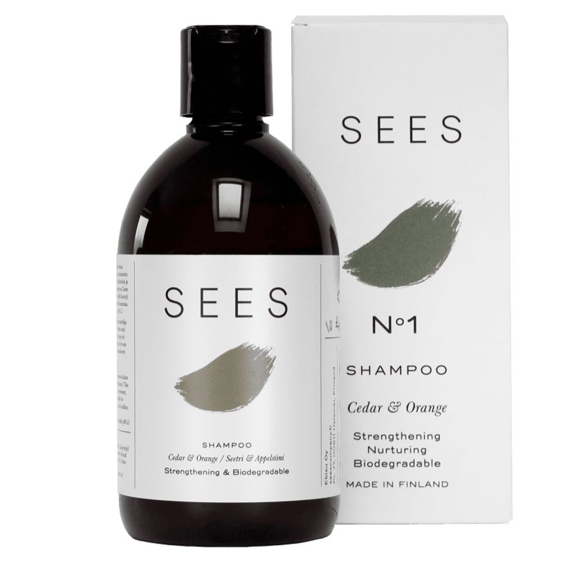 A bottle of SEES No1 Cedar & Orange Shampoo is shown next to its box. The cylindrical bottle is matte white with a black screw cap, and the rectangular box is also matte white with black text. Both display sans-serif typography. Label text includes “No1,” “Cedar & Orange,” and reference to biodegradable formula, strengthening, nurturing, and made in Finland. The background is plain and light-colored with no people or hands visible. Shot is front-facing at eye level.
