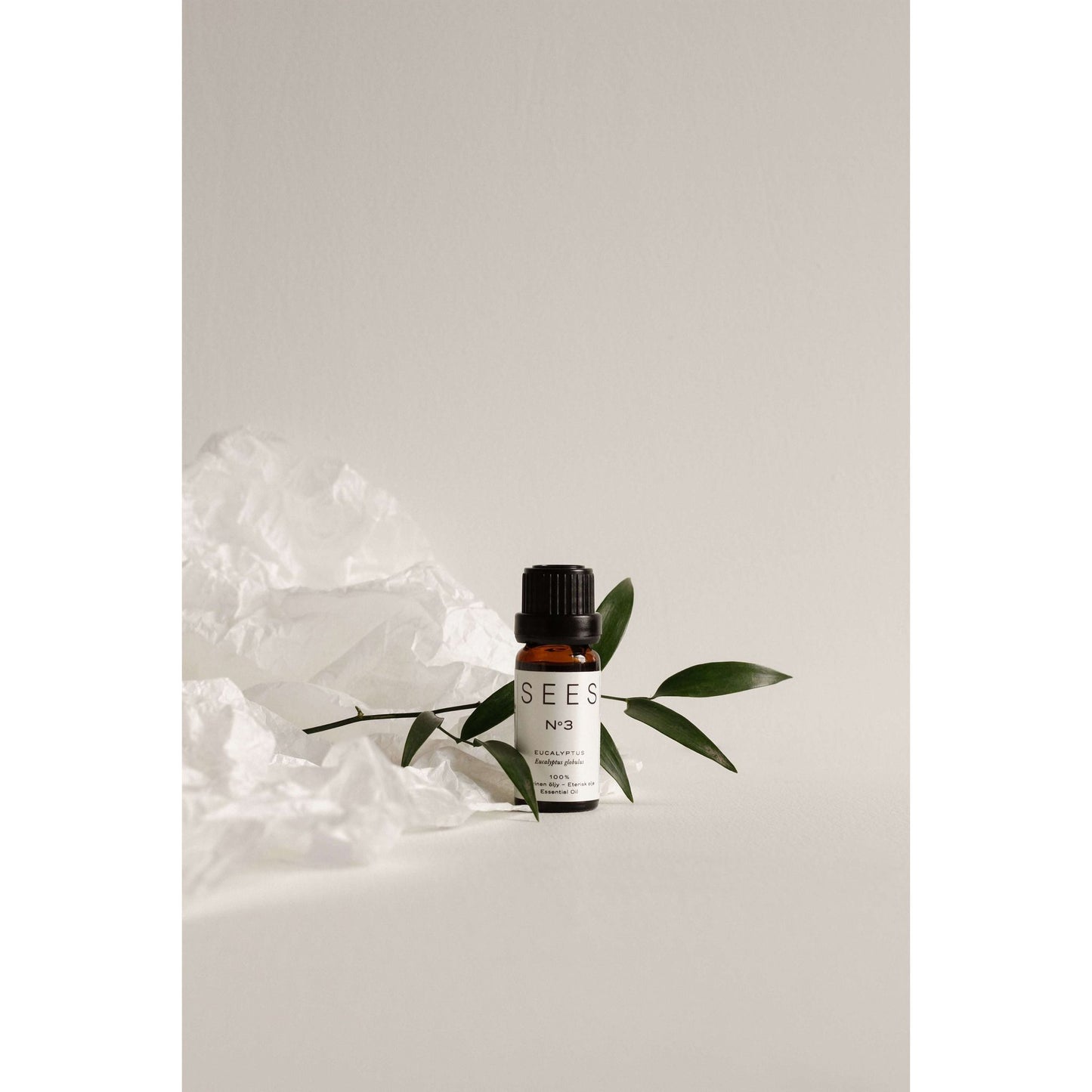 A small amber glass bottle labeled “SEES No3” stands upright. The bottle has a cylindrical shape with a screw cap. Black sans-serif text is printed on a white label. Green leaves are placed beside the bottle. Crumpled white tissue paper sits nearby. All items rest on a smooth, light-colored surface with a plain, uncluttered background in soft tones.