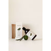 A bottle of SEES shampoo lies next to its rectangular box, both white with black sans-serif text. The bottle is cylindrical with a pump dispenser. A sprig of greenery and a whole lime are placed beside the products on a smooth white surface. No people or hands visible. Shot is flat lay from above. A bottle of SEES shampoo lies next to its box, accompanied by a sprig of greenery and a fresh lime, all beautifully arranged on a clean white surface.