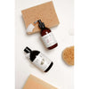 Two brown bottles of SEES skincare products rest on a white surface with beige bath sponges. The first bottle has a pump dispenser with soft foam visible at the opening; the second has a flip cap and small bubbles near the top. Both bottles have simple sans-serif SEES branding printed in white. Water droplets are present on the surface nearby, no people or hands appear in the image.