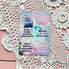 A postcard featuring Finnish motivational phrases on pastel watercolor splotches, displayed atop a white crochet doily and a pink wooden surface for inspiration. The postcard is rectangular and uncoated matte. Watercolor splotches include blue, green, yellow, and pink tones. Black sans-serif text in various weights is arranged diagonally and vertically within the colored splotches. No packaging or people are visible. Photographed from above in flat lay style.