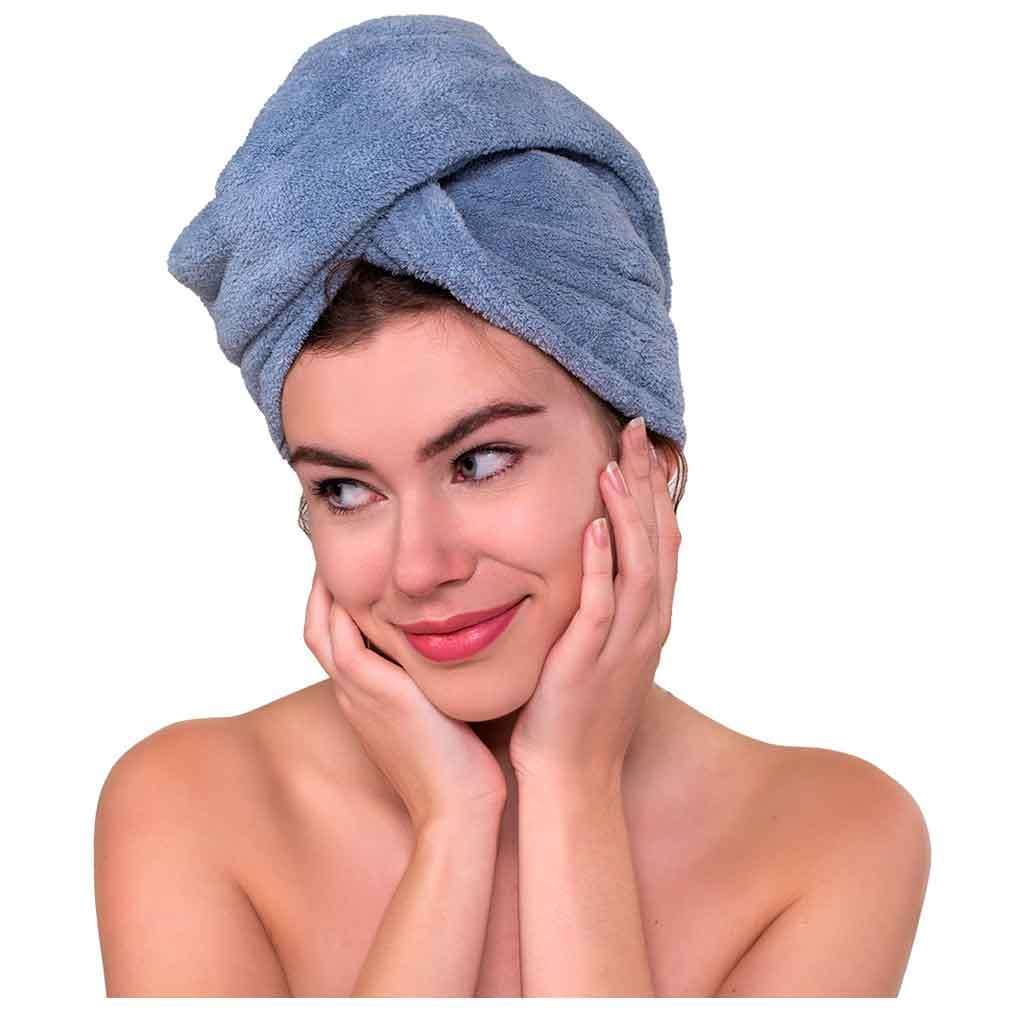 SkinMood Hair Turban Hiusturbaani; henkilö sinisellä pyyhkeellä hiuksissaan hymyilee pehmeästi, kädet kasvoillaan; olkapäät paljaina, katse hieman sivulle käännettynä; tasaisesti valaistu yhtenäinen valkoinen tausta ilman rekvisiittaa tai tekstiä; keskikokoinen lähikuva.