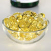 Swanson Vitamin D3 125 mikrog; pieni kirkas, pyöreä ja kiiltäväpintainen lasikulho täynnä läpikuultavia keltaisia pehmeäkapseleita; sileä, vaalea ja mattapintainen alusta; taustalla sumennettu suurempi pakkaus ilman luettavia etikettitietoja; ei ihmisiä tai käsiä.
