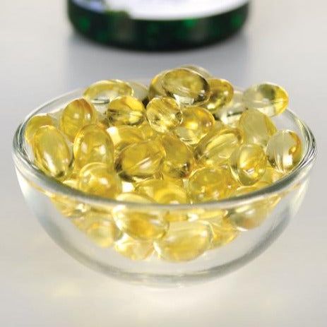 A small clear glass bowl, round and glossy, contains translucent yellow oval softgel capsules. The capsules fill the bowl. The surface beneath is smooth and light-colored with a matte finish. In the background, there is a larger container that is blurred; no readable label details are visible. No people or hands are present in the shot. A small clear glass bowl filled with translucent yellow softgel capsules, likely dietary supplements such as omega-3, sits on a light-colored surface. A blurred container