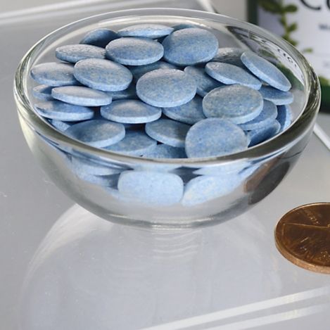 A clear glass bowl filled with round, flat blue tablets sits on a glossy glass surface beside a U.S. penny used for size comparison. The photo is shot from above. No visible packaging, labels, or text present. No people or hands in the image. The background is plain and uncluttered.