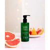 Taika Käsisaippua; vihreä muovinen pumppupullo tekstillä "Taika hand soap" seisoo pystyssä tasaisella valkoisella pinnalla; pullo lieriömäinen mattapinnalla, musta pumppuannostelija; taustalla viipaloitua greippiä ja appelsiinia; tausta pehmeästi valaistu ja lämminsävyinen, ei ihmisiä tai käsiä kuvassa.