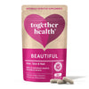Together Health Beautiful Hair, Skin & Nail Daily; vaaleanpunainen ja beige pussi hiusten, ihon ja kynsien hyvinvoinnille, pystyssä kahden beigen kapselin kera edessä; pussi mattapintainen ja suorakulmainen suljetulla yläosalla, selkeä lohkomainen teksti kollageenin tuotannosta; näkyvissä teksti "60 capsules"; vaalea, hyvin valaistu tausta, ei ihmisiä tai käsiä kuvassa.