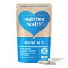 A blue and beige matte-finish pouch stands upright, with “together health BONE AID” printed in sans-serif type and additional text listing plant-based calcium, K2, D3, and 72 minerals. Three solid white capsules are placed directly in front of the pouch. The shot is on a plain background with no people or props visible.