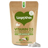 Brown resealable pouch labeled Together Vitamin D3 with coconut oil, featuring vegan and plant source icons. Matte finish, flexible packaging stands upright. Legible text in sans-serif font; illustrations of hazelnuts and green leaves are present on the front. Three white capsules placed directly in front of the pouch on a plain background. No people or hands are visible.