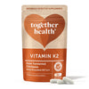 Together Health Vitamin K2; pakkaus kolmen valkoisen kapselin kera edessä; uudelleensuljettava pussi mattapintaisena ruskeana suorakulmaisena pyöristetyin kulmin; tuotenimi ja -tiedot painettu valkoisella ja keltaisella groteskikirjasimella; ei ihmisiä, käsiä tai rekvisiittaa, vaalea tausta; pakkaus sisältää 30 kapselia fermentoiduista kikherneistä, korostaen hyvää imeytymistä ja luonnollisia ainesosia.