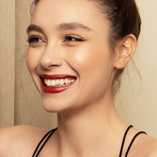 A woman with fair skin and dark hair in a bun smiles broadly. She wears red lipstick, natural makeup, and a black strap top. The background is neutral and softly lit, creating a timeless look. No visible product packaging or labels. No objects or props present in the image. Shot is a close-up portrait from the shoulders up.