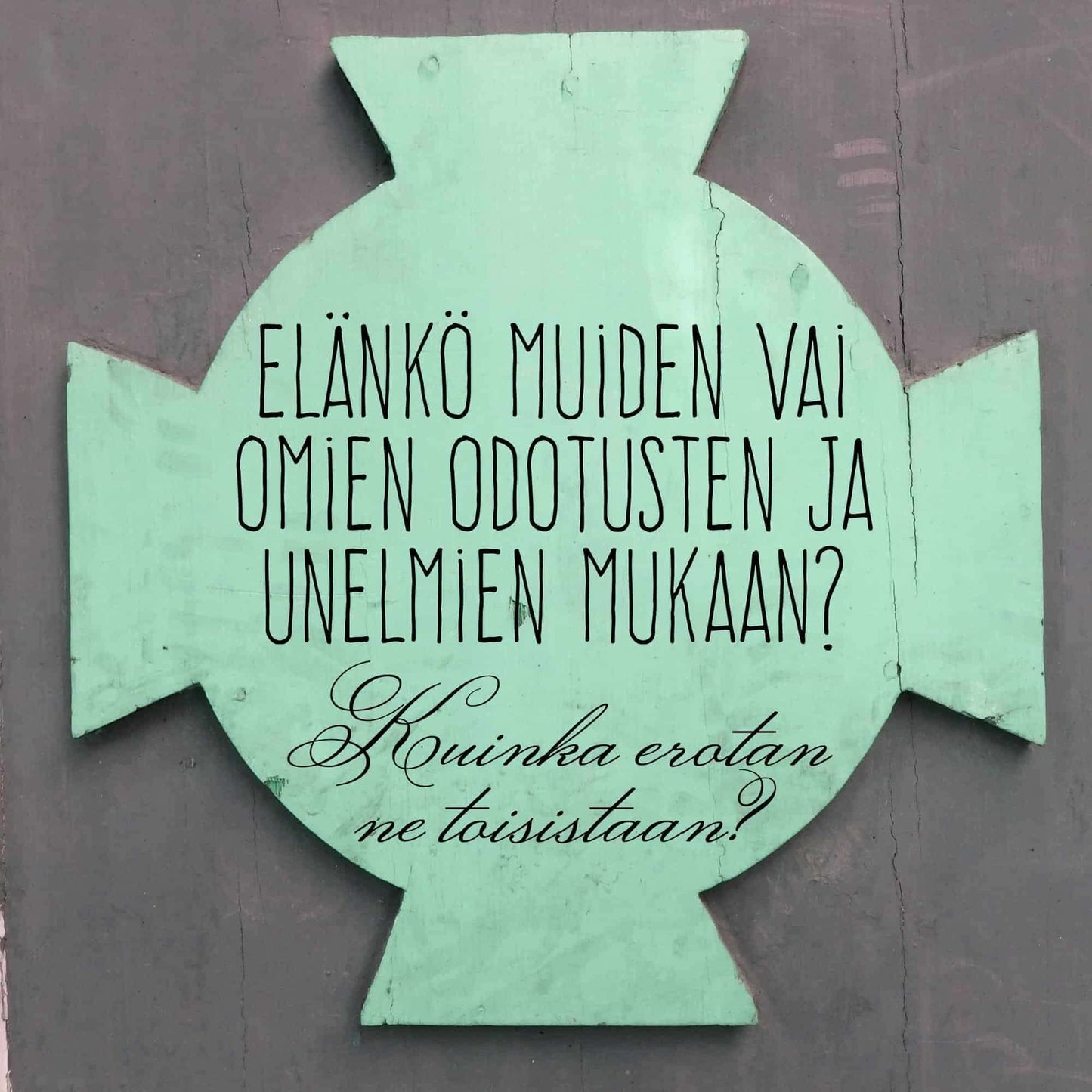Hidasta Elämää Voimauttavat Kysymykset -kortit; vihreä, ristinmuotoinen kyltti suomenkielisin tekstein pohtien, seuraako ihminen muiden vai omia unelmiaan ja miten tunnistaa ero; kyltin pinta mattainen valkoisin groteskikirjaimin; tausta yhtenäisen harmaa; ei ihmisiä tai toimintaa kuvassa; kuva näyttää tuotteen keskitettynä kehykseen.