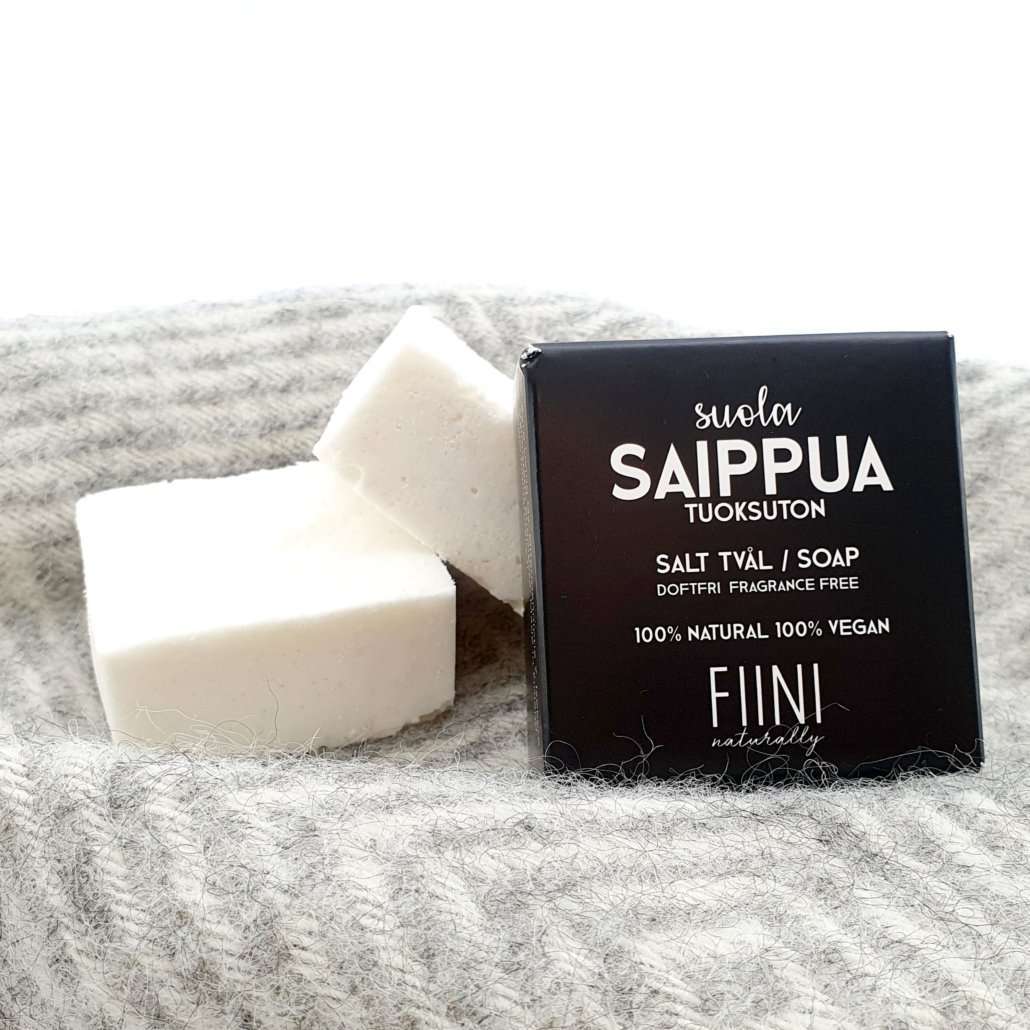 Fiini Naturally Suolasaippua Tuoksuton; kaksi suorakulmaista valkoista saippuapalaa mattapintaisina ja sileäreunaisina, vierellä musta laatikko tekstillä "Suola Saippua" lihavoidulla groteskikirjasimella; pakkaustekstissä mainitaan suolapohjaisuus, tuoksuttomuus, 100-prosenttinen luonnollisuus, vegaanisuus ja sopivuus kaikille ihotyypeille; kaikki tuotteet tekstuurisella vaaleanharmaalla alustalla; ei ihmisiä tai käsiä näkyvissä.