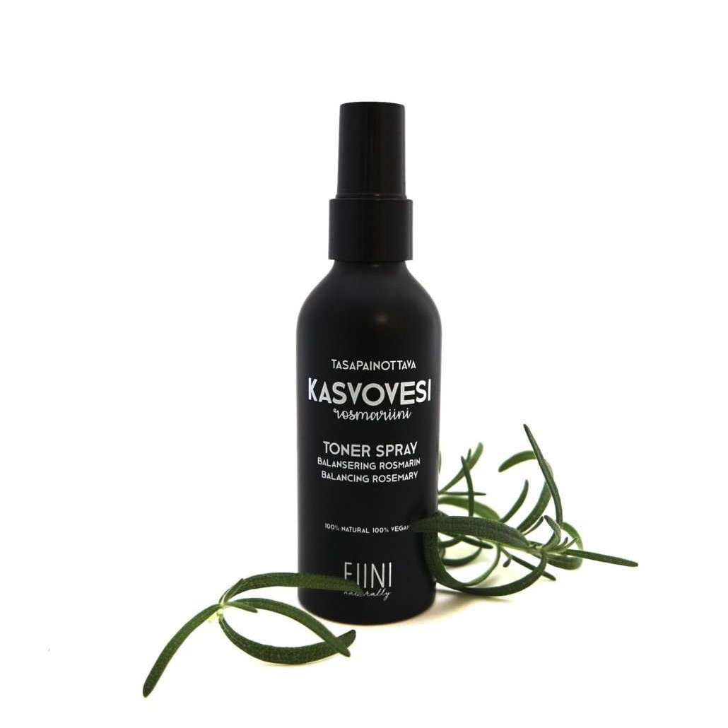 Fiini Naturally Tasapainottava Rosmariini Kasvovesi; musta pullo suihkusuuttimella, vierellä rosmariinioksia valkoisella taustalla; pullo sylinterinmuotoinen mattapintainen, suutin muovinen; etiketissä luettavissa teksti "Fiini Kasvovesi"; kuva otettu ylhäältä, näkyvissä vain tuote ja kasvi ilman ihmisiä tai käsiä.