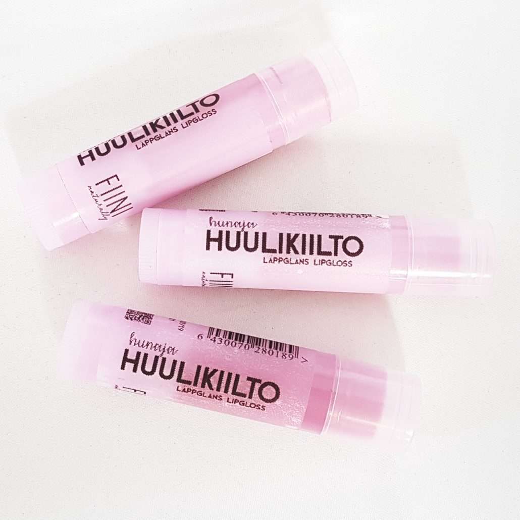 Fiini Naturally Hunajainen Huulikiilto; kolme huulikiiltotuubia aseteltuna valkoiselle alustalle; sylinterinmuotoinen kirkas muovipakkaus kiiltäväpintaisena, vaaleanpunaiset mattapintaiset korkit ja pohjat; etiketeissä suomen- ja ruotsinkielistä tekstiä sekä viivakoodit; tuotenimi lihavoidulla groteskikirjasimella; ei ihmisiä tai toimintaa, lähikuva.