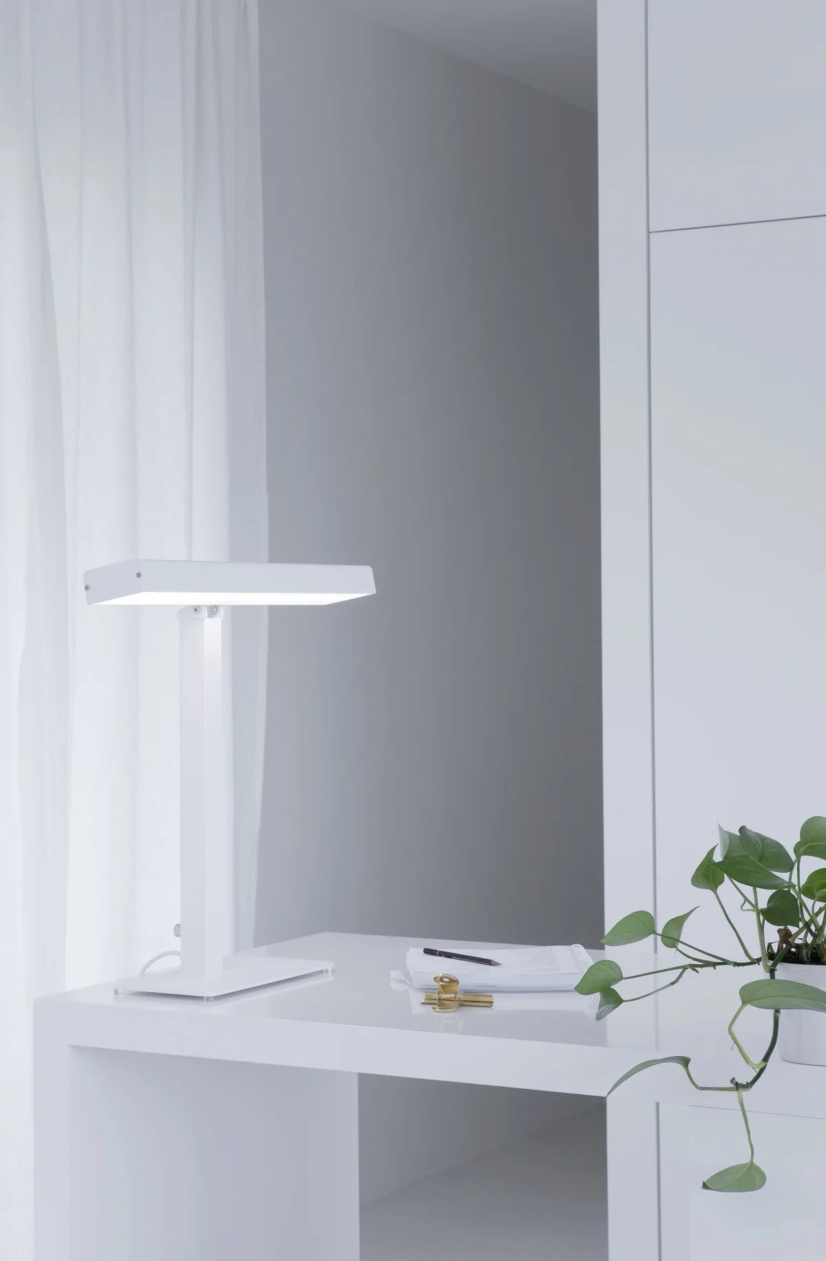Innolux Valovoima Pöytävalaisin; minimalistinen valkoinen pöytä modernin valkoisen lampun kanssa, papereita, kynä ja kultainen paperiliitin; vihreä lehtevä kasvi oikealla puolella; läpikuultavat valkoiset ikkunaverhot valoisassa, siistilinjaisessa huoneessa; mattapintaiset pinnat, ei ihmisiä tai käsiä; ei luettavaa etikettitekstiä; yläkuva luonnonvalossa.