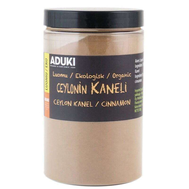 Aduki Luomu Ceylon Kaneli; lieriömäinen kirkas muovipurkki täynnä hienoksi jauhettua vaaleanruskeaa kanelia, suljettu mustalla mattapintaisella kierrekannella; etuetiketti painettu useilla kielillä, näyttää Aduki-brändin ja tekstin "Ceylon cinnamon" lihavoidulla groteskifontilla; ei ihmisiä tai käsiä näkyvissä; neutraali, tasainen tausta; kuva suoraviivainen tuotteen lähikuva.