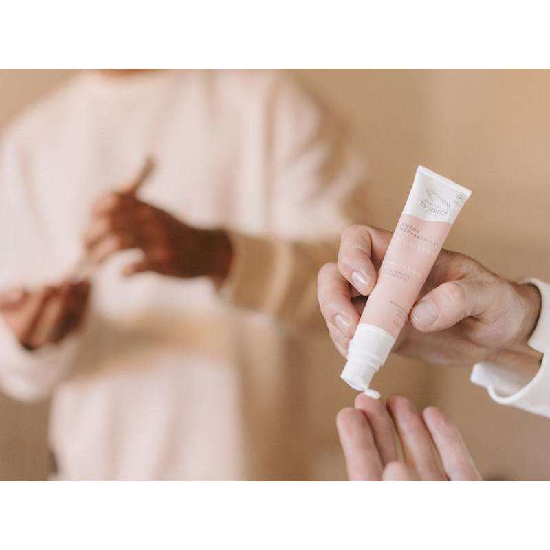 Alga Cicosa Reparative Cream Korjaava Hoitovoide; henkilö puristaa voidetta vaaleanpunaisesta, mattapintaisesta tuubista sormelleen, taustalla toinen henkilö pitelee sivellintä; tuubi soikea ja muovinen ilman luettavaa etikettitekstiä; molemmat henkilöt vaaleissa yläosissa; ympäristö pehmeästi valaistu, todennäköisesti sisätiloissa, neutraali tausta; lähikuva keskittyy käsiin ja tuotteisiin.