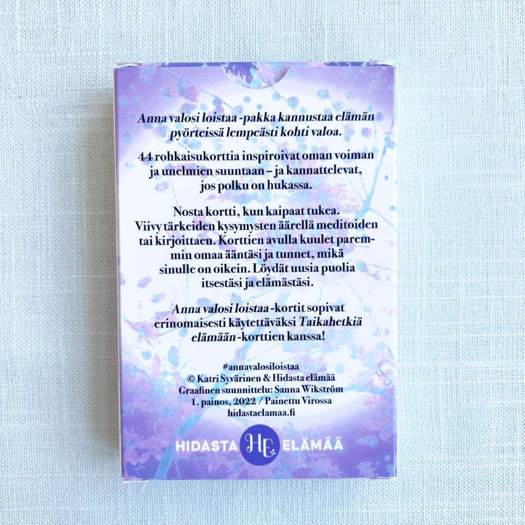 Hidasta Elämää Anna valosi loistaa -rohkaisukortit; pastellisävyinen, suorakulmainen ja mattapintainen korttipakan pakkaus violetein ja sinisin vesiväriläikin sekä Hidasta Elämää -logo; suomenkielinen groteskiteksti kertoo hyvinvointia ja mielentyyneyttä tukevista rohkaisukorteista; pakkaus pystyssä valkoisella taustalla.