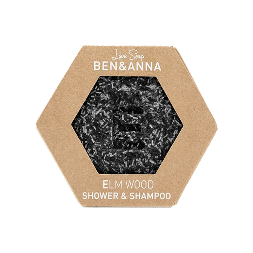 Ben & Anna Shower & Shampoo palasaippua Elm Wood; kuusikulmainen, ruskea ja mattapintainen pahvipakkaus terävin reunoin, ikkunasta näkyy tumma ja tekstuurinen saippua; valkoinen versaaliteksti pakkauksen etusivulla; vaalea tausta.