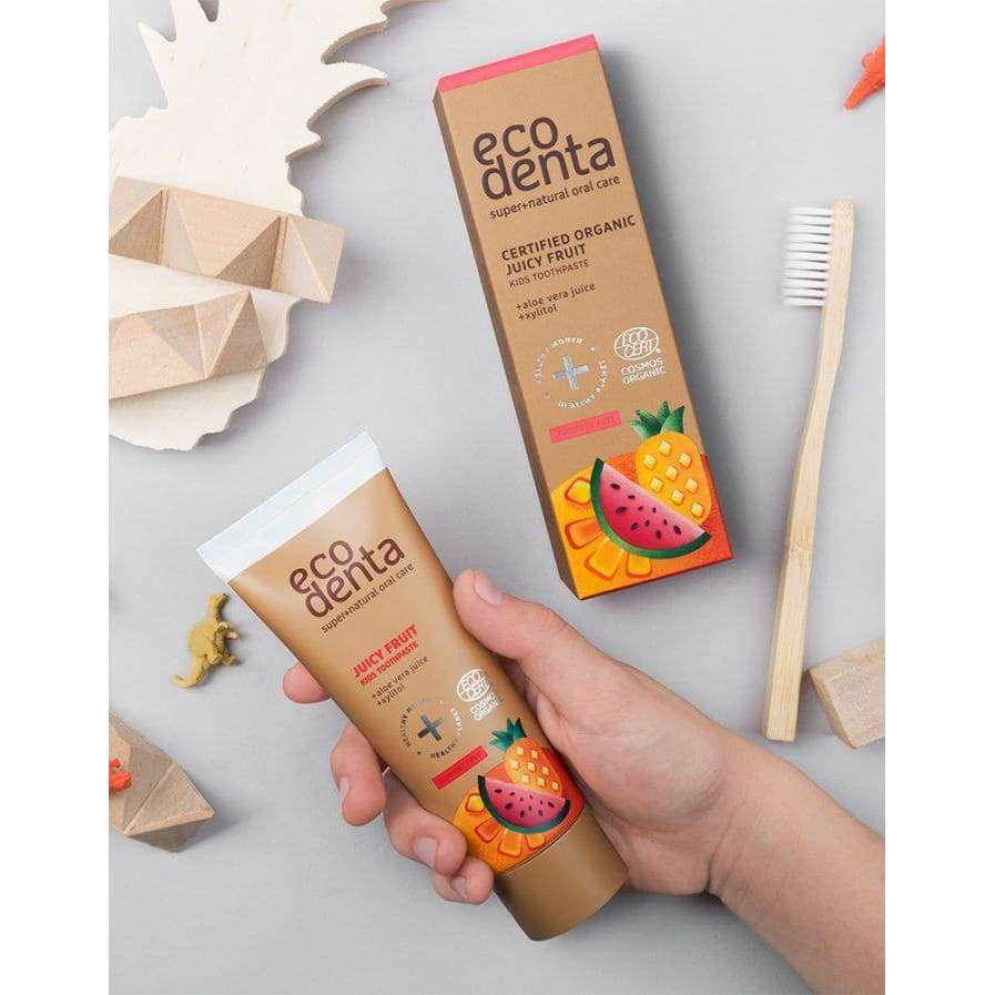 Käsi pitelee sylinterimäistä, pehmeäpintaista tuubia, jossa lukee "Ecodenta Certified Organic Juicy Fruit toothpaste" lihavoiduin kirjaimin; hammastahnatuubi ja vastaava suorakulmainen pakkaus koristeltu kiiltävin värikkäin hedelmäkuvituksin; lähellä kaksi puista lelufiguuria ja muovinen hammasharja mattaharmaalla pinnalla; lähikuva, jossa vain osa kädestä näkyy.
