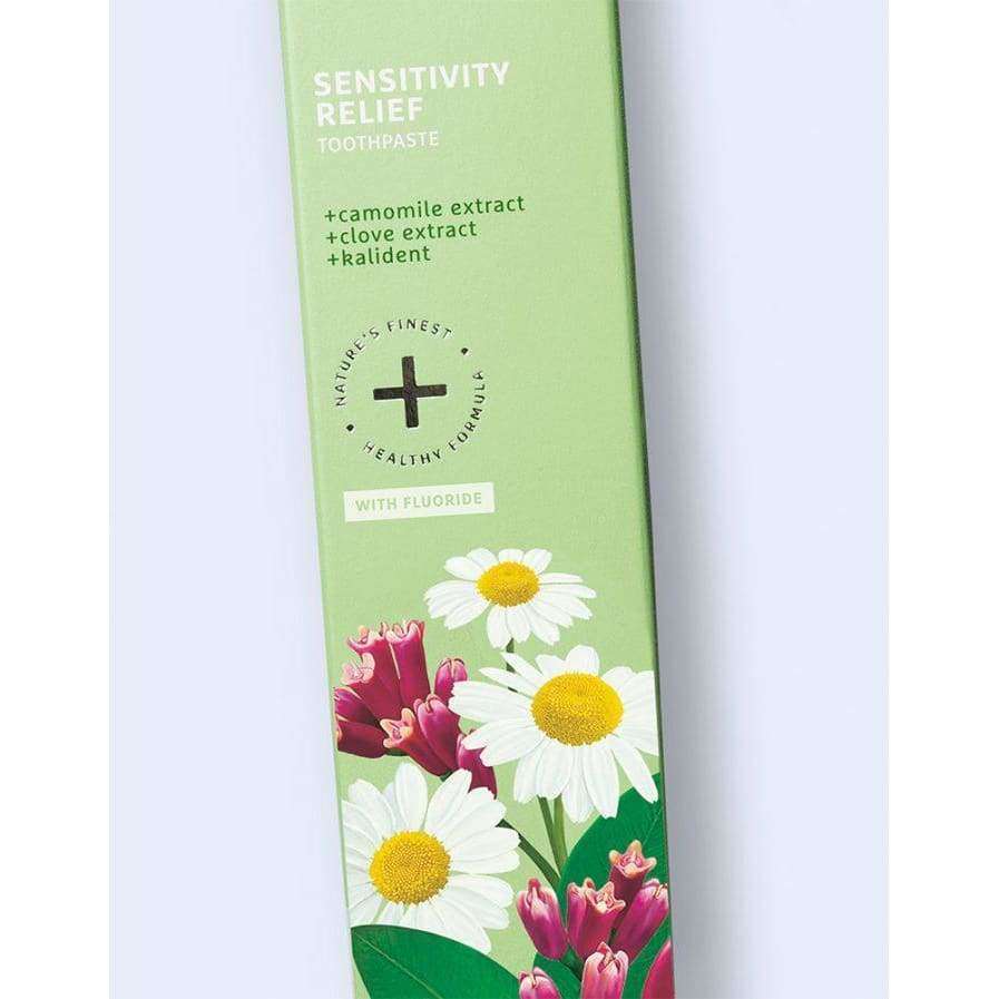 Ecodenta Sensitivity Relief Herkkien Hampaiden Fluorihammastahna; vihreä hammastahnarasia tekstillä "Sensitivity Relief" kamomilla- ja neilikkauutteella, kamomillankukka- ja neilikkakuvituksin sekä tekstillä, joka mainitsee fluorin ja hellävaraisen koostumuksen; rasia suorakulmainen mattapinnalla; typografia lihavoitua groteskia pääetiketissä; ei ihmisiä tai käsiä näkyvissä; tausta valkoinen, studiotyylinen tuotekuva.