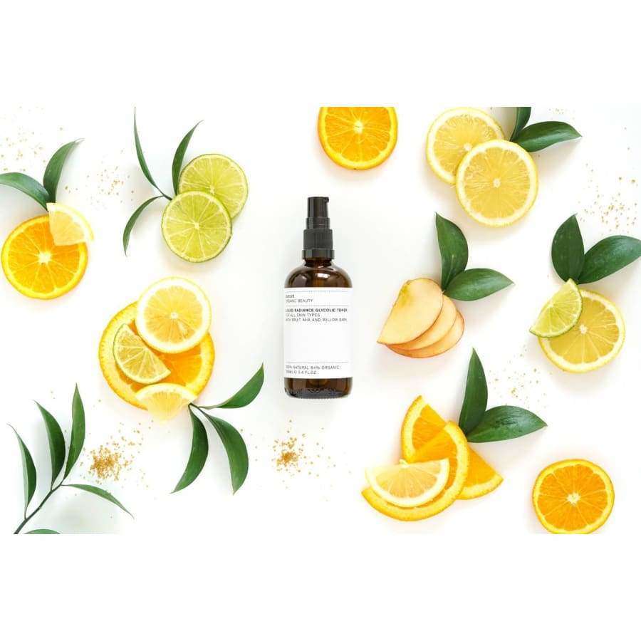 Evolve Liquid Radiance Glycolic Kasvovesi; ruskea suihkepullo kiiltäväpintaisena, lieriömäinen muoto, todennäköisesti lasia tai muovia, pystyssä; musta suihkesuutin, ei näkyvää etikettiä tai tekstiä pullossa; ympärillä appelsiini-, lime- ja omenaviipaleita sekä tuoreita vihreitä lehtiä; kaikki aseteltu yhtenäiselle valkoiselle taustalle tasaisessa valaistuksessa.