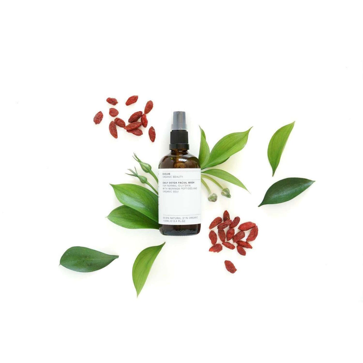 Evolve Daily Detox Facial Wash Puhdistusgeeli -suihkepullo vihreiden lehtien ja punaisten marjatertujen ympäröimänä valkoisella taustalla; ruskea, sylinterimäinen ja mattapintainen pullo valkoisella muovisella suihkusuuttimella; etiketin teksti ei ole selvästi luettavissa kuvassa; kuvattu ylhäältä päin, ei ihmisiä näkyvissä.