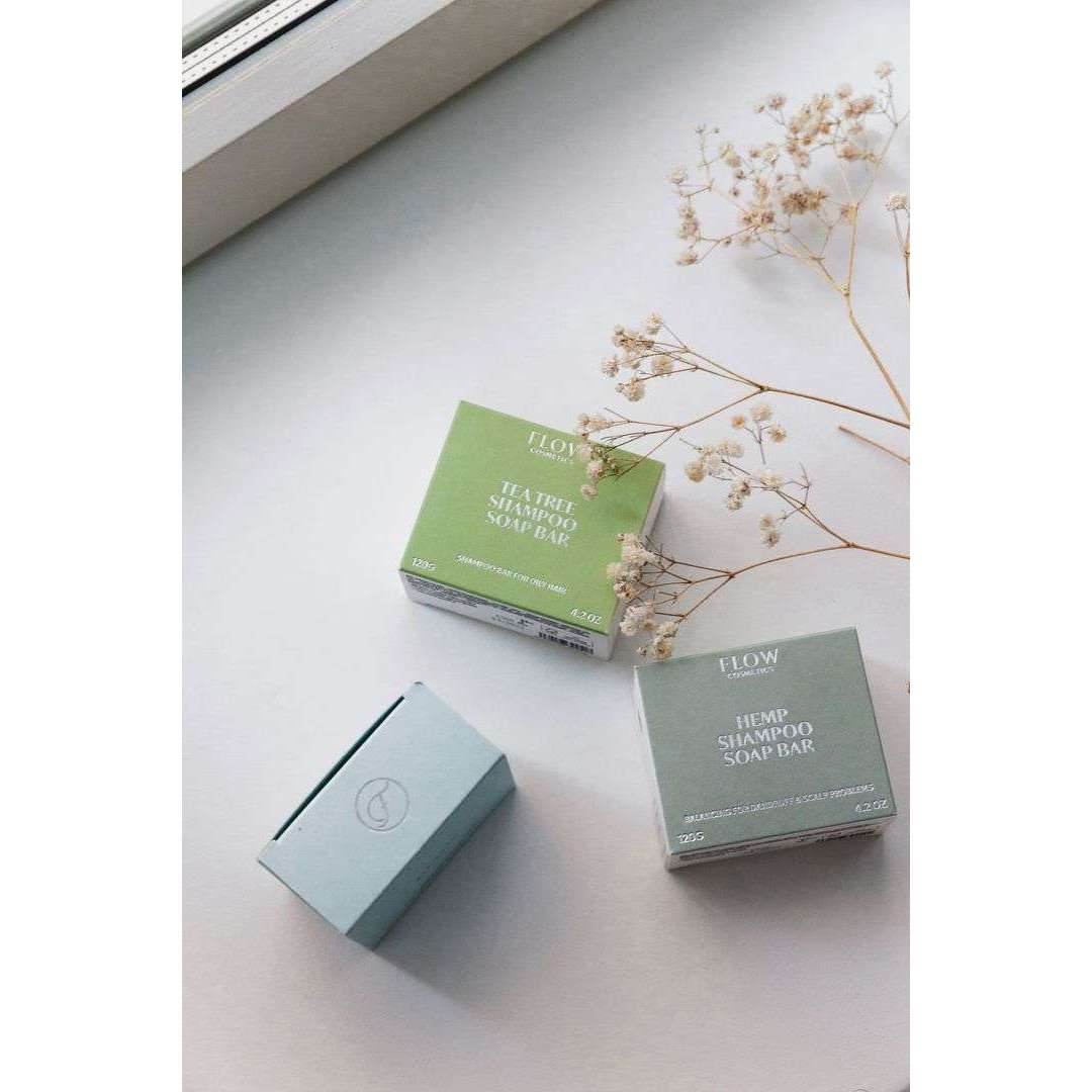 Kolme pastellinsävyistä, suorakulmaista rasiaa vaalealla, tasaisella alustalla; kahdessa rasiassa näkyy teksti "Shampoo Soap Bar" selkeällä groteskityypillä; kaikki rasiat mattapintaista pahvia; vieressä kuivattuja valkoisia kukkia; kuva otettu ikkunan lähellä voimakkaassa päivänvalossa; ei ihmisiä tai käsiä.