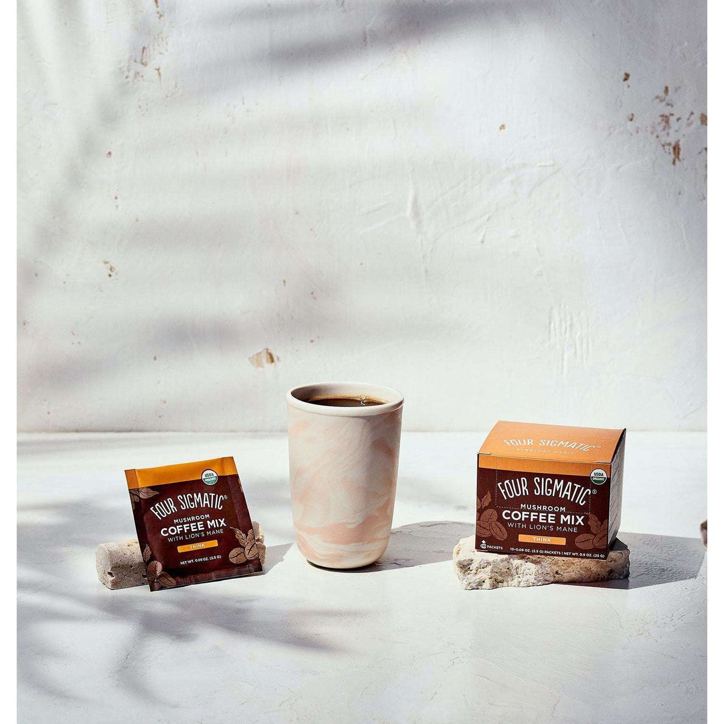 Four Sigmatic Mushroom Coffee Mix Lions Mane; Four Sigmatic Mushroom Coffee Mix -pussi, keraaminen kuppi täynnä kahvia sekä Four Sigmatic-kahvirasia aseteltuna mattavalkoiselle pinnalle pehmeässä luonnonvalossa; pussissa ja rasiassa Four Sigmatic-logo selkeällä lihavoidulla typografialla; ei ihmisiä tai käsiä näkyvissä; sienipitoinen sekoitus korostuu asetelmassa.