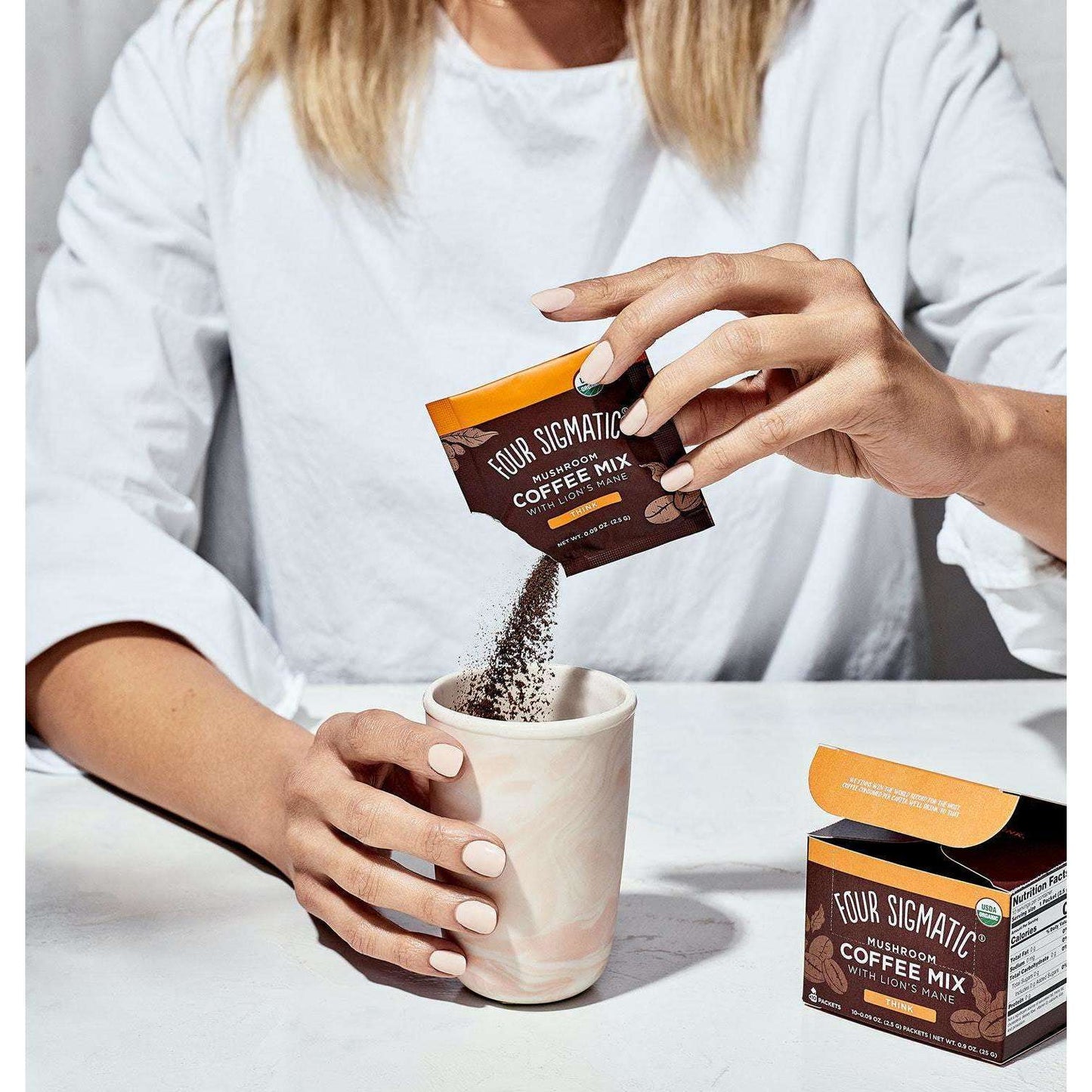 Four Sigmatic Mushroom Coffee Mix Lions Mane; henkilö kaataa Four Sigmatic-pikakahvisekoitusta suorakulmaisesta pussista valkoiseen keraamiseen mukiin, jossa tummanharmaa marmorikuvio ja pyöreä muoto; rasia tekstillä "Adaptogen Coffee Mix" mattaruskeine pakkauksineen ja selkeine groteskitekstein lepää läheisellä tasaisella pöytätasolla; tausta neutraali, pinta sileä, tuotebrändaus kameraan päin.